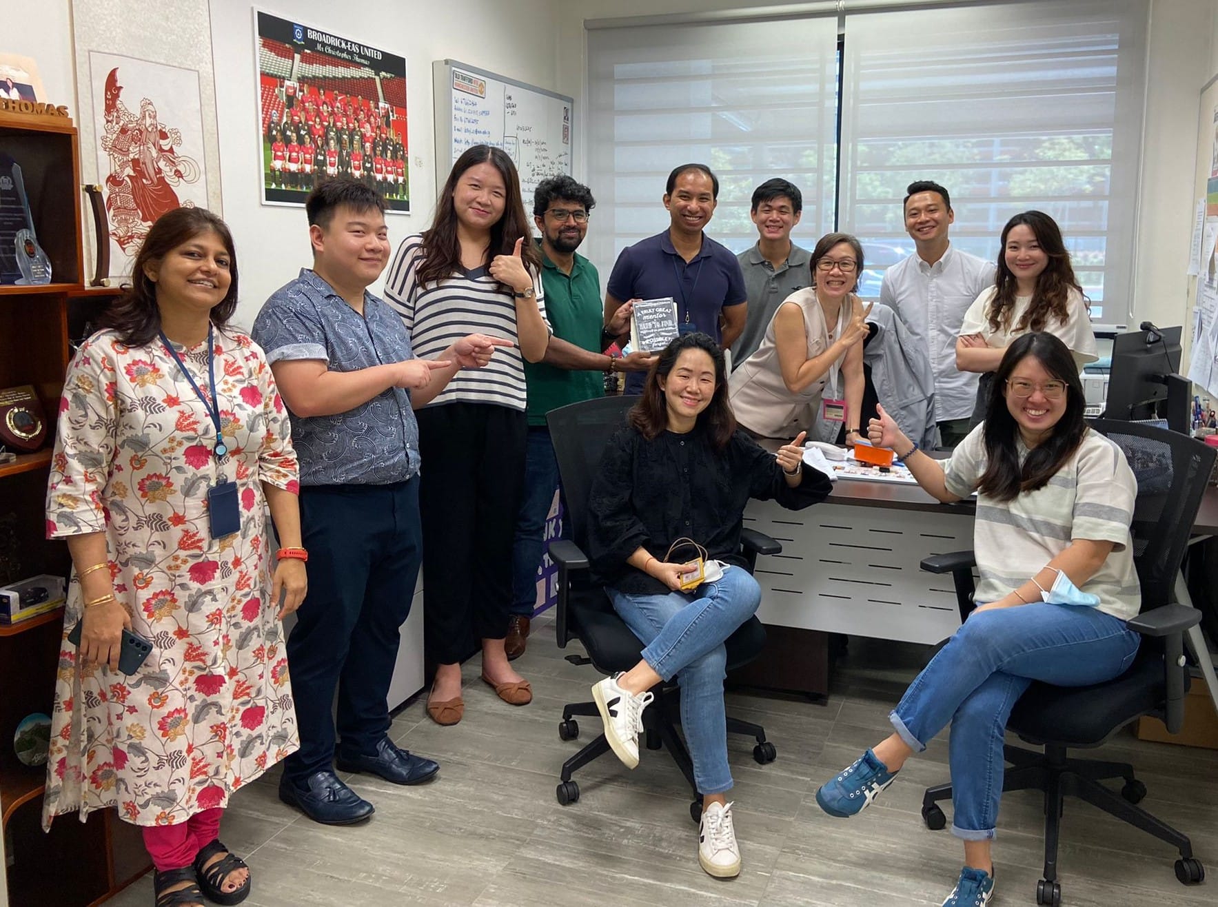 A group of eleven smiling colleagues posing together in an office, some seated and some standing, with posters on the wall.